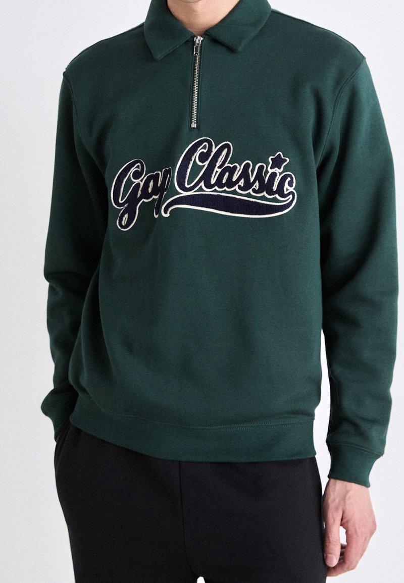 Pull-over vert foncé avec une fermeture éclair quart, avec le texte "Gay Classic" brodé en cursive sur la poitrine, porté par une personne aux mains relâchées.