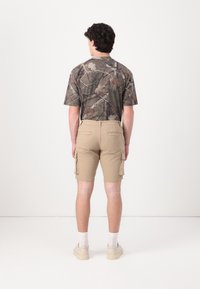T-shirt camouflage à manches courtes avec des nuances de vert et de marron ; shorts cargo beiges avec poches latérales et silhouette ajustée, portés avec des baskets.