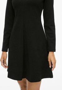 Czarna sukienka z długim rękawem, dopasowaną górą i rozkloszowanym dołem, wykonana z miękkiej tkaniny. Posiada okrągły dekolt i prosty brzeg.