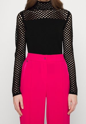 Femme portant un haut noir à manches longues en maille avec des découpes circulaires et un col montant, associé à un pantalon taille haute rose vif.