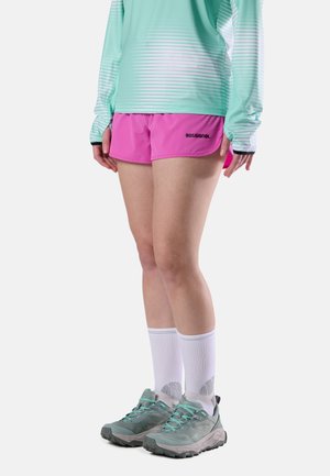 Persona con camiseta de manga larga verde menta, pantalones cortos rosa brillante Rossignol, calcetines blancos hasta la mitad de la pantorrilla y zapatos de senderismo gris, de pie sobre fondo blanco.