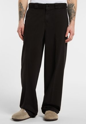 Uomo che indossa pantaloni neri larghi, pantofole beige e una canotta bianca, con tatuaggi visibili su entrambi gli avambracci.