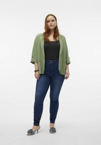 Veste légère verte sur un haut noir, jeans skinny bleus et mules à talons gris avec des accents floraux. Texture lisse et design ajusté.