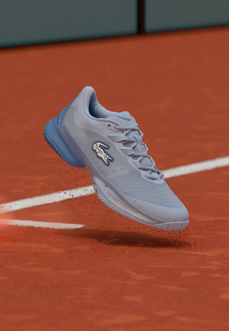 Ανοιχτό μπλε αθλητικό παπούτσι τένις με υφασμάτινο επάνω μέρος, σχέδιο με κορδόνια και εμφανές λογότυπο Lacoste στο πλάι, σε κίνηση πάνω από γήπεδο με χώμα.