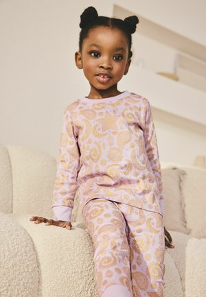 Młoda dziewczyna z dwoma kokami, ubrana w jasnoróżową piżamę z wzorami złotych gwiazd i księżyców, siedząca na teksturowanej beżowej sofie w pomieszczeniu.