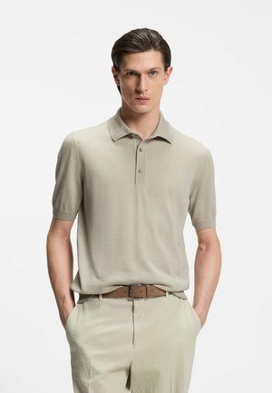 Homme portant un polo à manches courtes beige clair et un pantalon beige texturé avec une ceinture marron, debout les mains dans les poches devant un fond blanc.