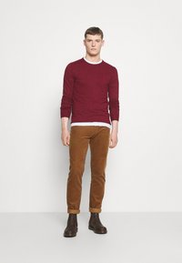 Burgunderfarbene Langarmbluse über einem weißen Rundhalsausschnitt, kombiniert mit braunen Cordhosen und dunkelbraunen Stiefeletten. Glatte Textur, taillierte Passform.