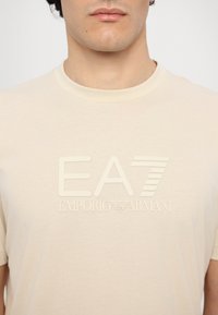 Šviesiai rudas trumpomis rankovėmis marškinėliai su iškiliu logotipu "EA7 EMPORIO ARMANI" priekyje. Minkšta tekstūra su apvalia kaklo iškirpte.