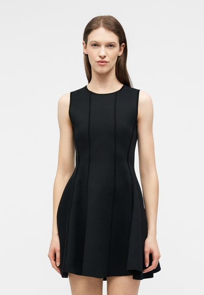 Γυναίκα με μακριά καστανά μαλλιά που φοράει μαύρο εφαρμοστό φόρεμα χωρίς μανίκια με κάθετες ραφές, στέκεται μπροστά σε απλό φωτεινό φόντο.