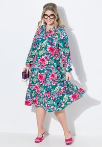 Vestido floral en verde azulado con patrones rosas y verdes, de mangas largas y cintura ceñida. Combinado con sandalias de tacón rosas y un bolso morado.