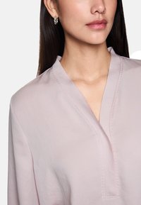 Light pink blouse with a split neckline, soft texture, and subtle stitching detail visible along the edges. Model wears silver hoop earrings.