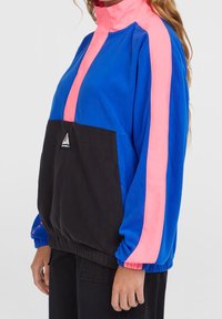 Personne portant une veste en polaire color block bleu, noir et rose avec un col montant et des poignets élastiques.