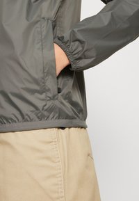 Gray, lightweight jacket with elastic cuffs, featuring a side pocket and a smooth, shiny texture, paired with beige pants.