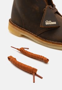 Braune Lederstiefel mit einer hellbraunen Gummisohle, versehen mit einem dreieckigen Anhänger. Zwei passende rostfarbene Schnürsenkel liegen daneben.