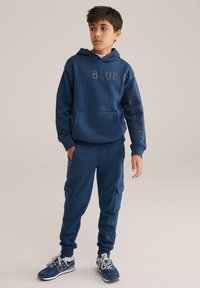 Mörkblå hoodie med texten "BLUE", kombinerad med matchande joggers med sidofickor. Slät yta, avslappnad design och åtsittande mudd.