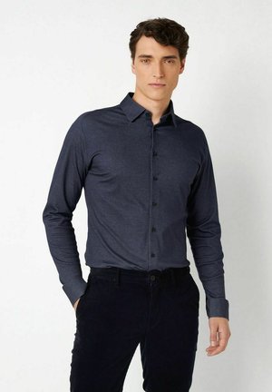 Junger Mann mit dunklen lockigen Haaren, der ein eng anliegendes, langärmeliges dunkelblaues Hemd mit Knöpfen und schwarze Hose trägt, vor einem schlichten weißen Hintergrund steht.