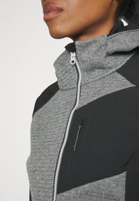 Veste zippée grise et noire avec un col montant, tissu gris texturé, panneaux noirs lisses et une petite poche zippée à l'avant.