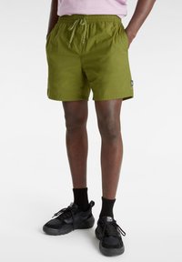 Pantalones cortos verdes con cinturilla elástica y cordón, que presentan bolsillos laterales y una etiqueta en el lado izquierdo. Llevados con zapatillas negras y calcetines.