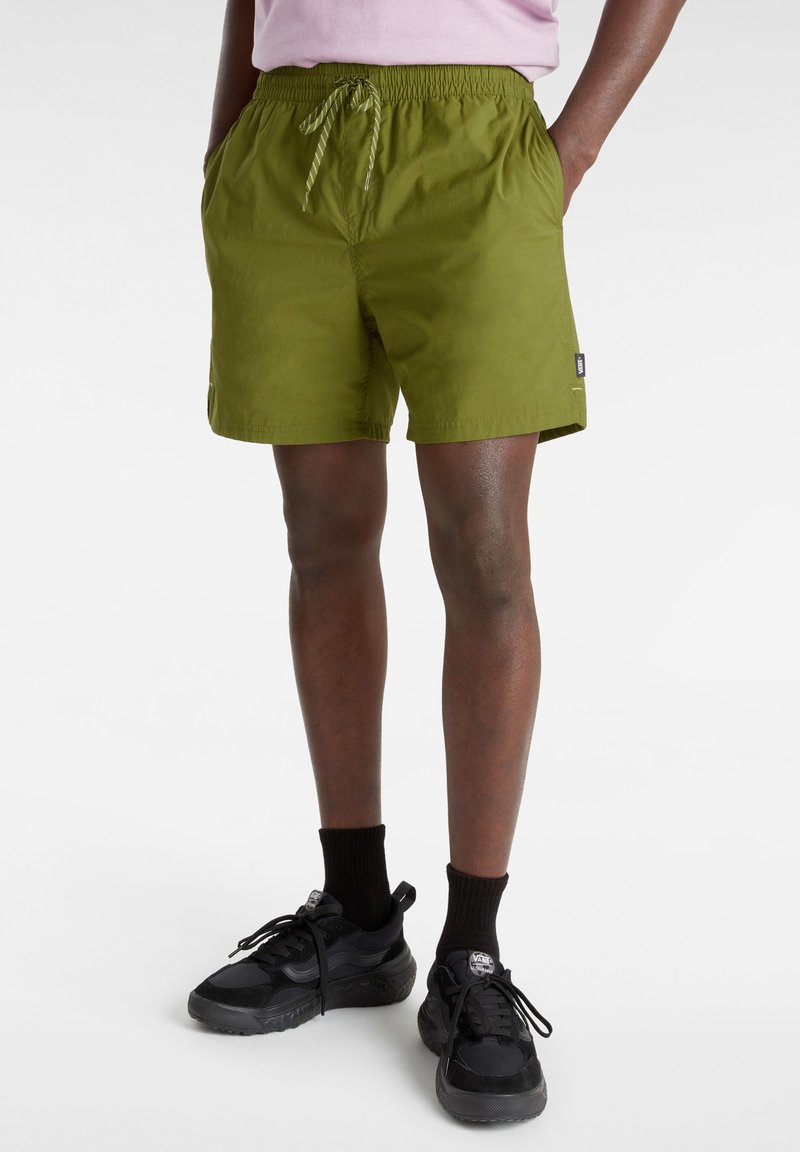 Pantalones cortos verdes con cinturilla elástica y cordón, que presentan bolsillos laterales y una etiqueta en el lado izquierdo. Llevados con zapatillas negras y calcetines.