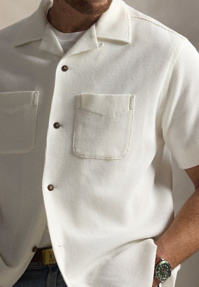 Λευκό πουκάμισο με κοντά μανίκια και υφή υφάσματος, δύο τσέπες στο στήθος και καφέ κουμπιά. Φορεμένο πάνω από λευκή μπλούζα, συνδυασμένο με τζιν.