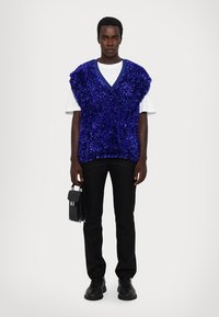 Ryškiai mėlynas be rankovių megztinis su pūkuota tekstūra, dėvimas ant balto marškinėlio, kartu su juodomis kelnėmis ir mažomis juodomis rankinėmis.