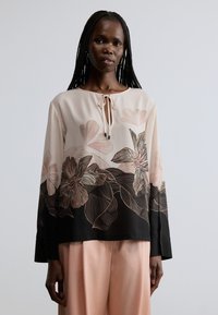 Blouse beige clair avec imprimés floraux en rose et noir, dotée d'un col rond, d'un détail noué et de longues manches. Texture de tissu lisse.