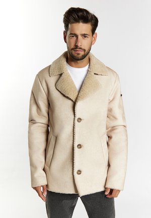 Uomo che indossa una giacca beige in shearling con grande colletto e bottoni sopra una camicia bianca e pantaloni scuri, in piedi davanti a uno sfondo bianco.