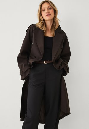 Blonde woman wearing a dark brown trench coat over a black top and high-waist black pants with a brown belt, standing against a light background.