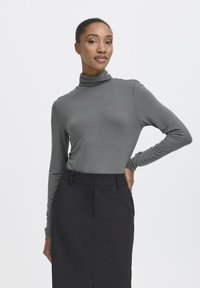 Donna con pelle scura e capelli raccolti in un bun, che indossa un maglione grigio a collo alto e una gonna nera, in piedi con una mano sui fianchi su uno sfondo neutro.