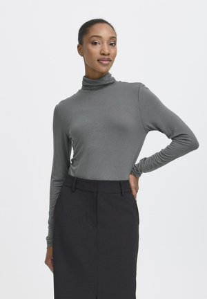 Donna con pelle scura e capelli raccolti in un bun, che indossa un maglione grigio a collo alto e una gonna nera, in piedi con una mano sui fianchi su uno sfondo neutro.