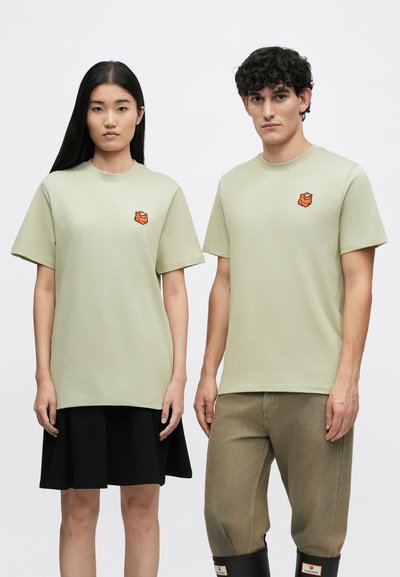 Svetlozelené bavlnené tričká s krátkymi rukávmi a malým oranžovým logom na hrudi, ktoré nosia dvaja jednotlivci stojaci vedľa seba.