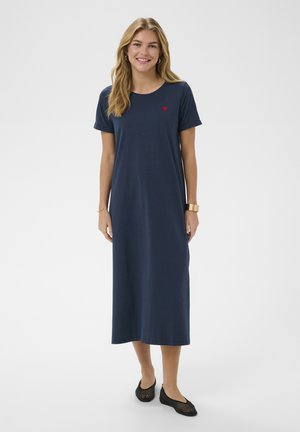 Femeie zâmbitoare cu păr blond, purtând o rochie midi albastru închis cu mâneci scurte și o inimă roșie mică pe piept, încălțată cu balerini negri din plasă.