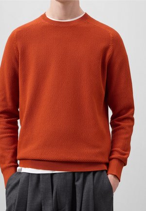 Mann trägt einen strukturierten orangen Pullover mit langen Ärmeln über einem weißen Hemd und grauen plissierten Hosen, Hände in den Taschen, vor hellem einfarbigem Hintergrund.