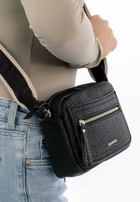 Black textured crossbody bag with a gold zipper, featuring a front pocket and a striped adjustable strap. Suitable for casual use.