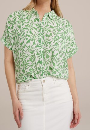 Femme portant une chemise boutonnée à motif de feuilles tropicales vertes et blanches, rentrée dans un jean blanc taille haute sur un fond uni.