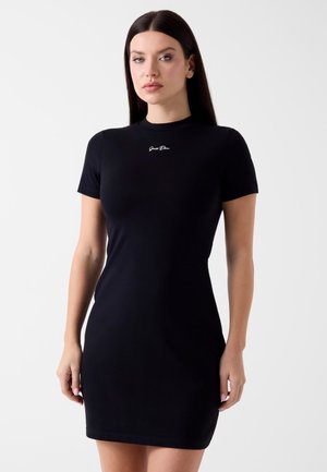 Femme aux longs cheveux foncés portant une robe noire ajustée à manches courtes avec une petite inscription blanche sur la poitrine, debout devant un fond clair et uni.