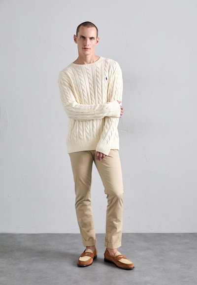 Cream cable-knit sweater with a round neckline, tan chinos, and brown loafers featuring a light cream accent. Simple, textured design.