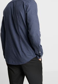 Laivastonsininen napillinen paita, jossa kaareva helma ja pitkät hihat, valmistettu teksturoidusta kankaasta, pidetty mustien housujen päällä. Yksinpainikkeiset kalvosimet.