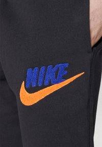 Pantaloni della tuta neri con un logo "NIKE" blu in rilievo e un design a swoosh arancione, entrambi con texture ricamata, e una tasca laterale.