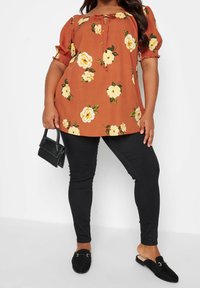 Bluzka w kolorze rdzy z żółtym kwiatowym wzorem, krótkie bufiaste rękawy i dekolt z wycięciem. Dopasowane czarne legginsy i czarne buty wsuwane.
