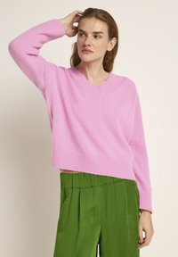 Rosafarbener, locker sitzender V-Ausschnitt-Pullover mit gerippten Manschetten und Saum, aus weichem Material, kombiniert mit grünen, gefalteten weiten Hosen.