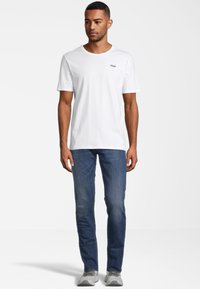 Weißes Baumwoll-T-Shirt mit kurzen Ärmeln, das ein kleines gedrucktes FILA-Logo auf der linken Brust trägt. Kombiniert mit blauen Jeans und grauen Turnschuhen.