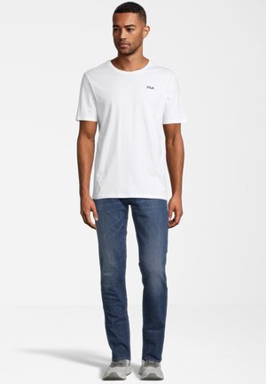 T-shirt bianco in cotone a maniche corte, con un piccolo logo FILA stampato sul lato sinistro del petto. Abbinato a jeans blu e sneakers grigie.
