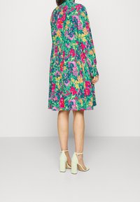 Blumendruckkleid in Türkis mit rosa, gelben und lila Blumen. Lange Ärmel, plissiertes Design, knielang, kombiniert mit hellgrünen High Heels.