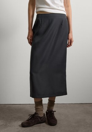 Persoon in een zwarte midi-rok, beige sokken, donkerbruine sneakers en een witte mouwloze top tegen een effen witte muur en een grijze vloer.