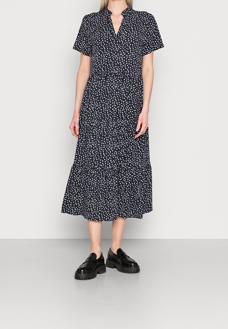 Femme portant une robe midi noire à pois blancs, manches courtes, et des chaussures compensées noires épaisses, debout contre un mur uni.
