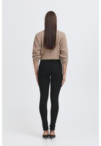 Jean skinny noir taille haute, texture lisse, avec deux poches arrière, associé à un pull marron clair. Modèle montré de dos.