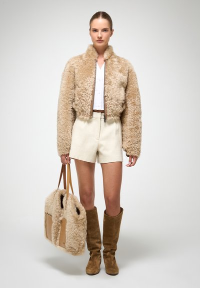 Béžová umelá kožušinová bomber bunda s vysokým golierom, krémové šortky, hnedé čižmy po kolená a zodpovedajúca chlpatá taška držiaca sa na ramenných popruhoch.