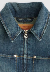 Γιακάς τζιν μπουφάν με ορατή πορτοκαλί ραφή, ασημένιο φερμουάρ και μέρος από τις μπροστινές τσέπες με μεταλλικά καρφιά.