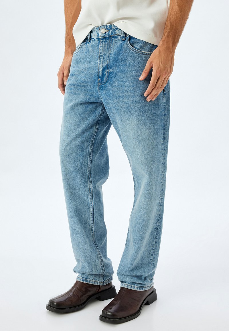 Jeans de mezclilla azul claro con un corte relajado, diseño de cinco bolsillos y costuras visibles. Combinados con botas de cuero marrón oscuro.
